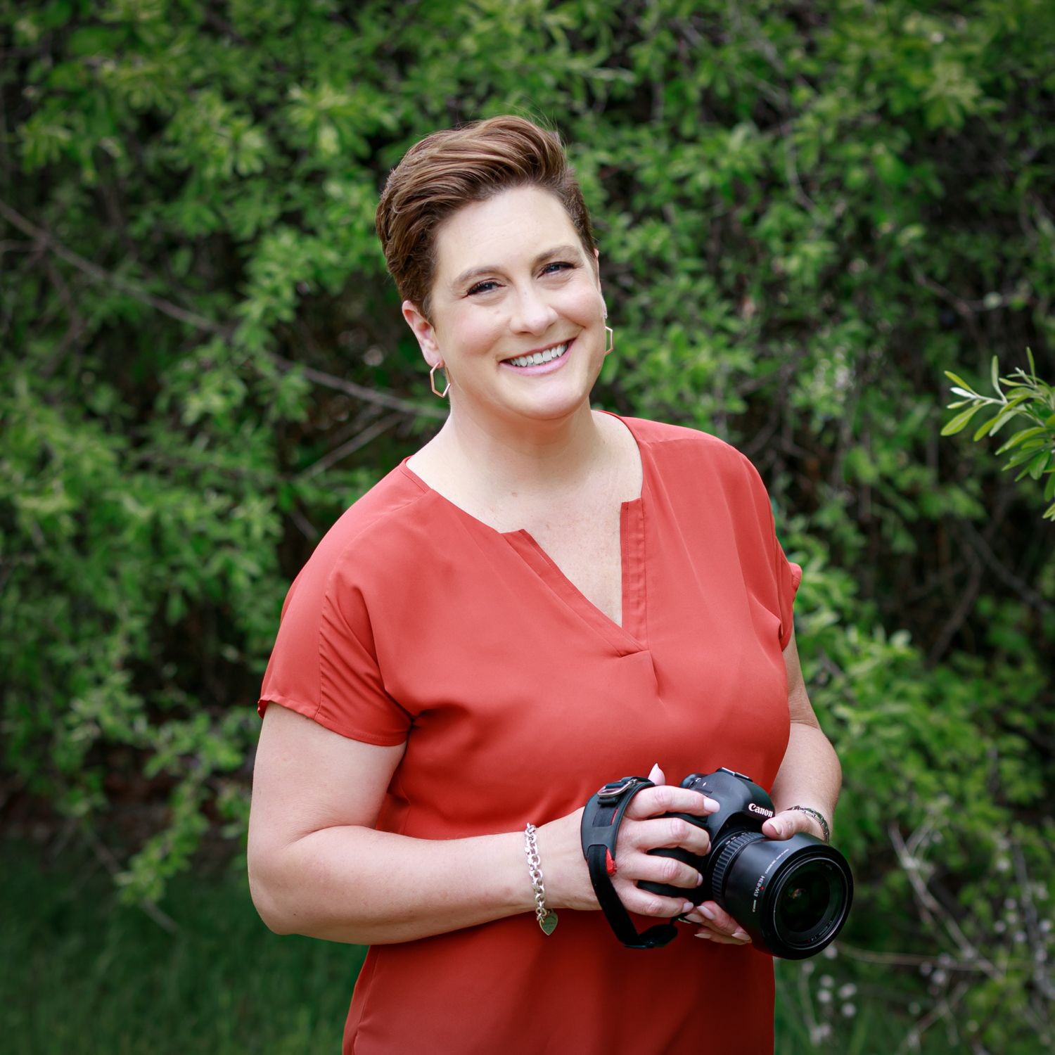 brunette with orange shirt holding camera with green trees in background