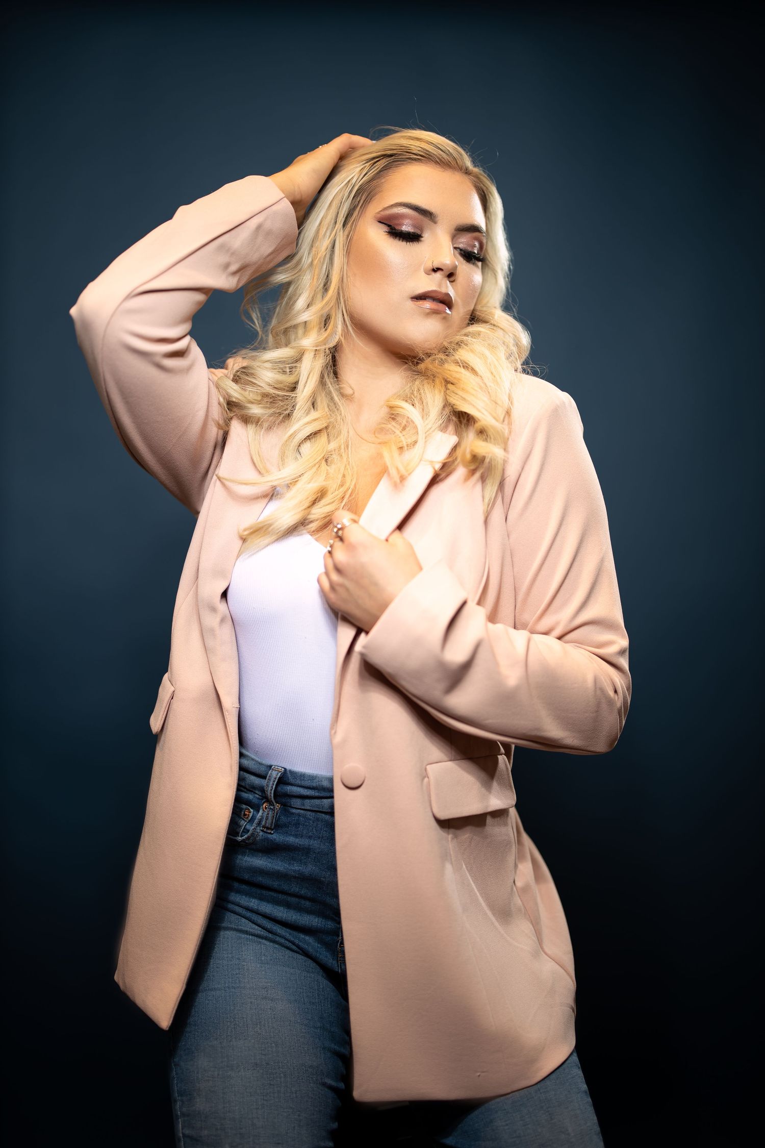 Soft glamour portrait in pink blazer and denim against dark background.
