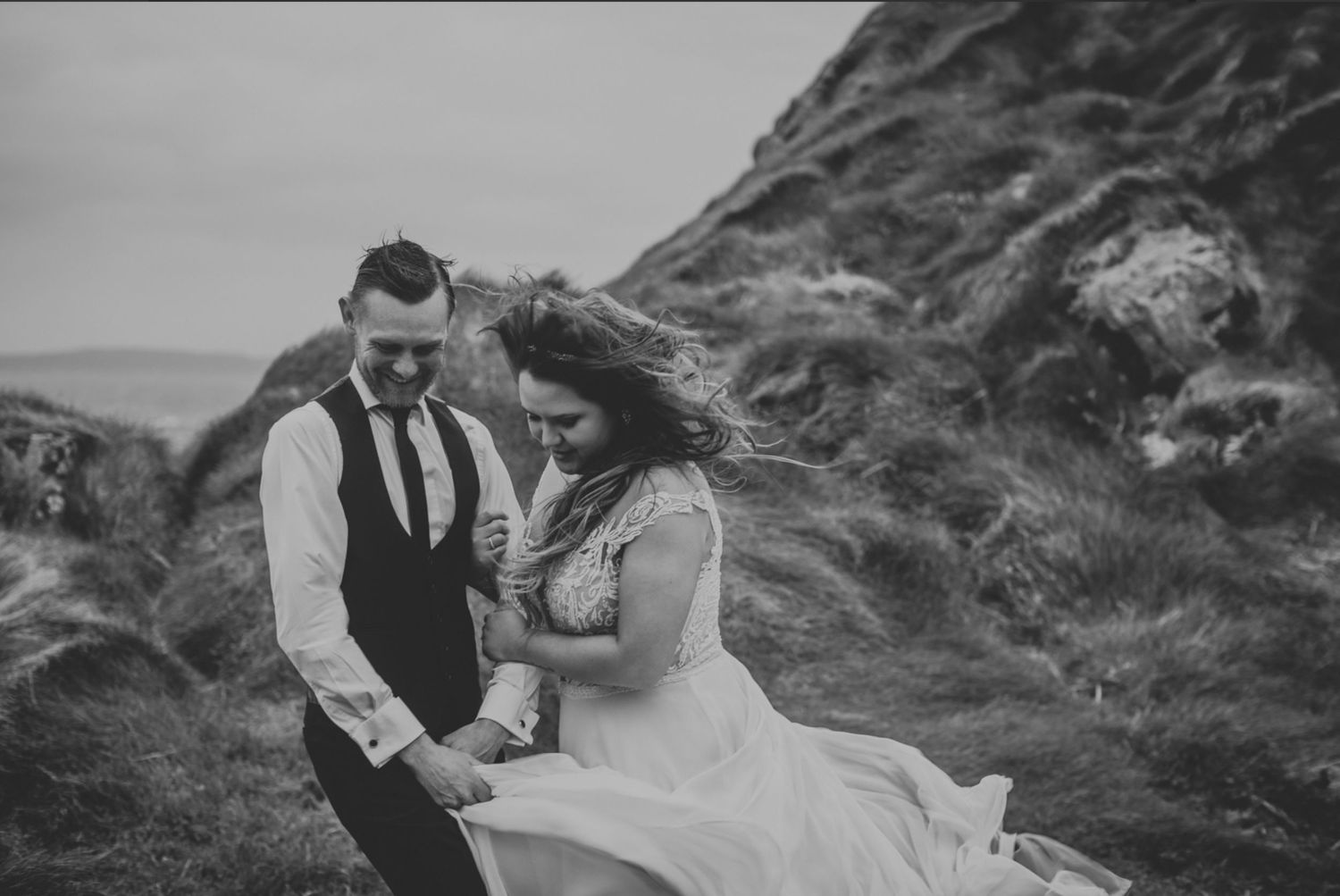 Couple shares an intimate moment on windswept coastal cliffs in black and white.