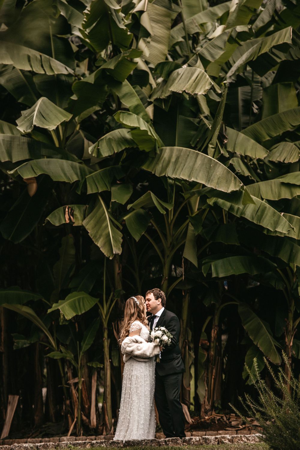 A romantic wedding moment captured beneath towering banana trees with lush green foliage.