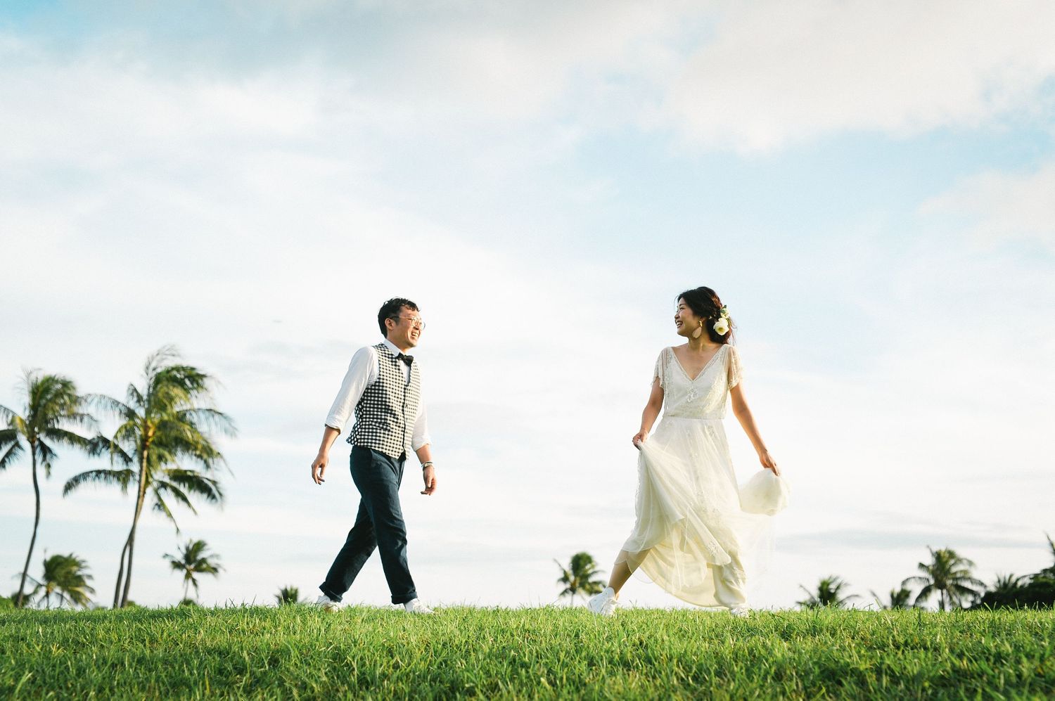 Happy bride and groom walking on lush green grass with tropical palm trees.