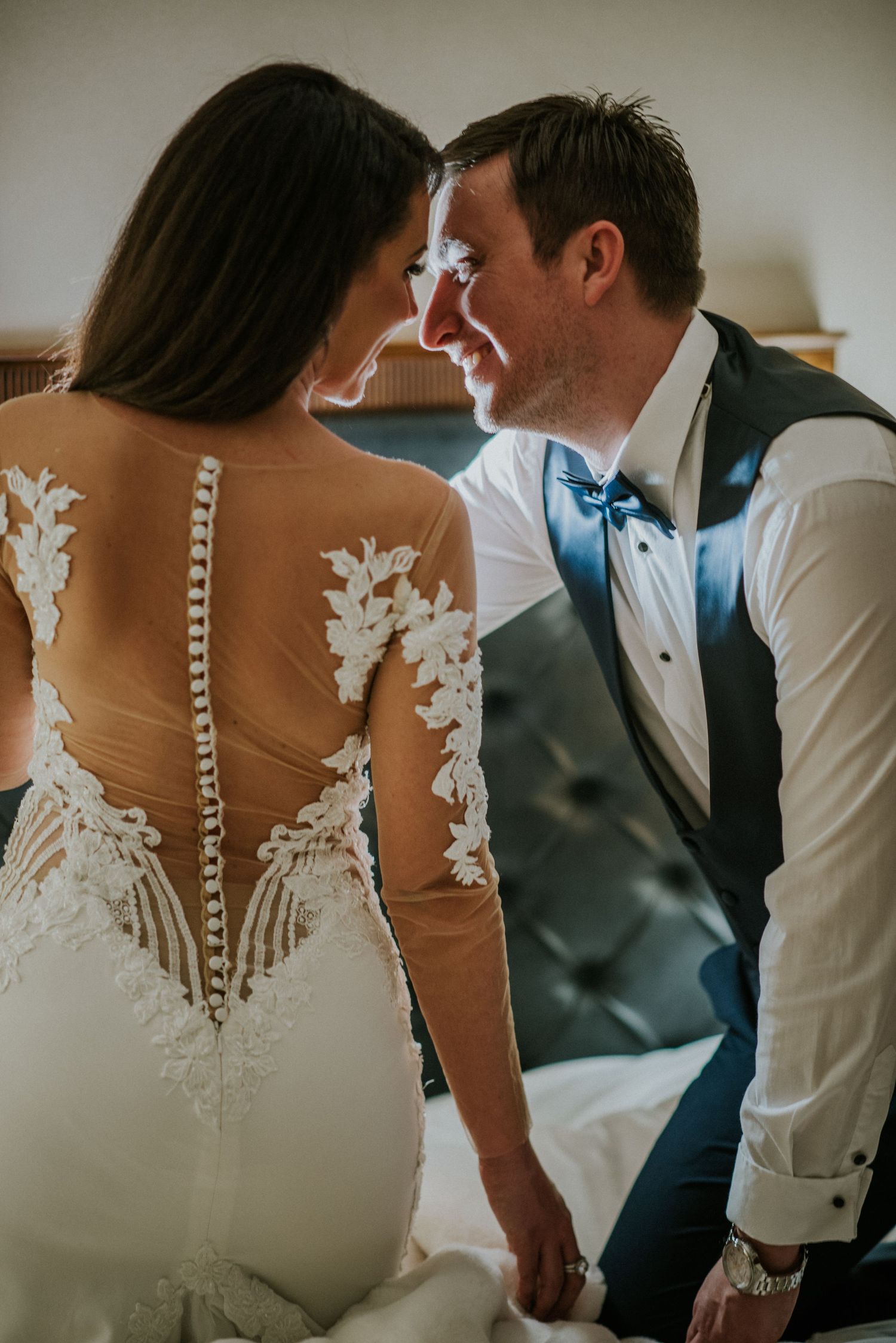 Close-up detail of a wedding dress with intricate lace appliques and sheer back as newlyweds share an intimate moment.