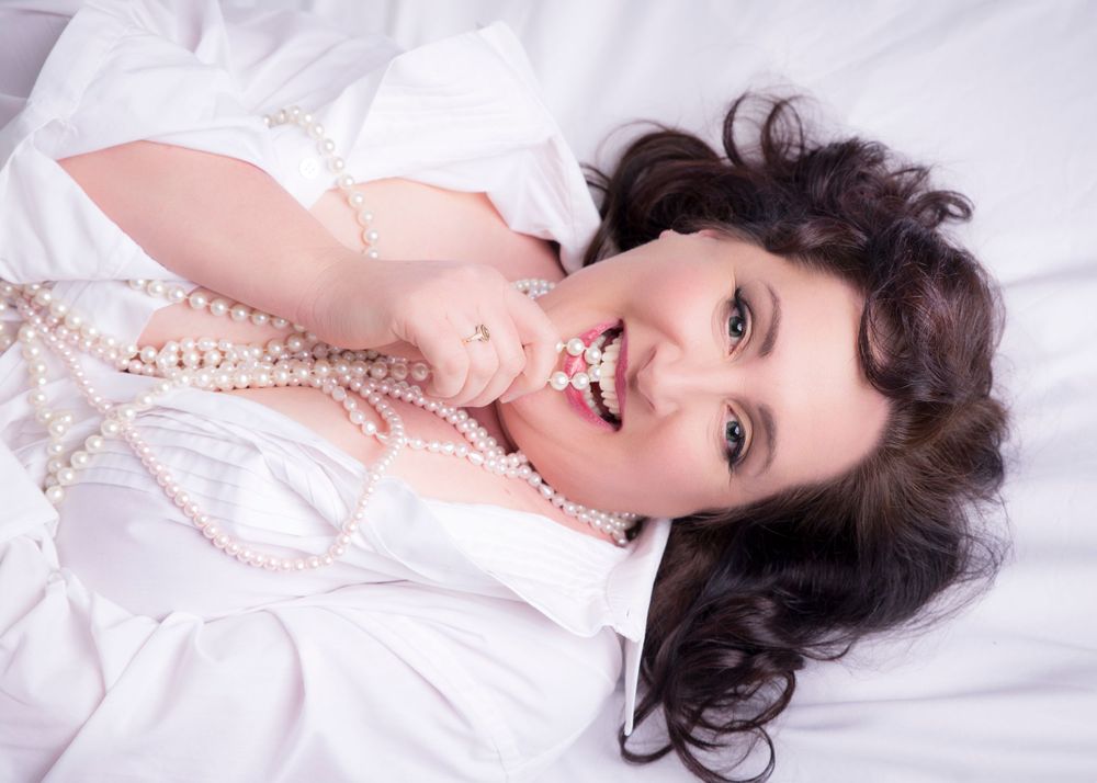 Woman lying on white bedding wearing pearls in vintage-inspired boudoir photo.
