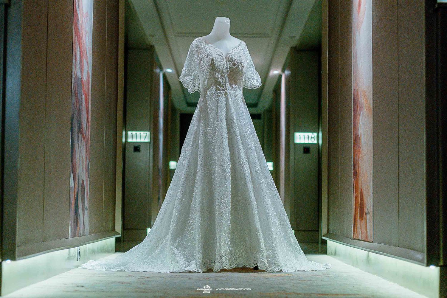 A white lace wedding dress on display in an elegant hallway with dramatic lighting.