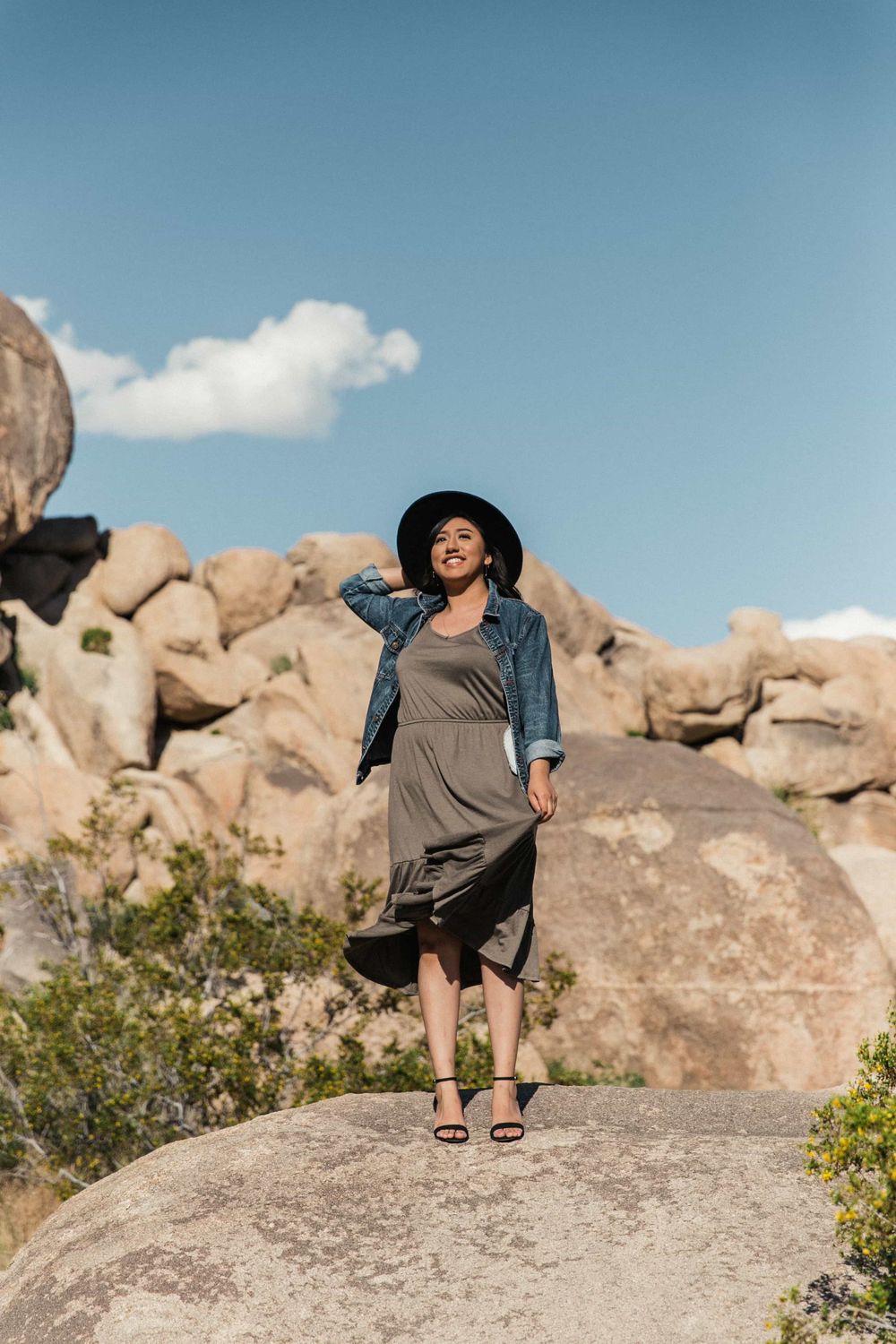 Stylish woman in gray dress, denim jacket, and black hat poses on boulder in rocky desert landscape.