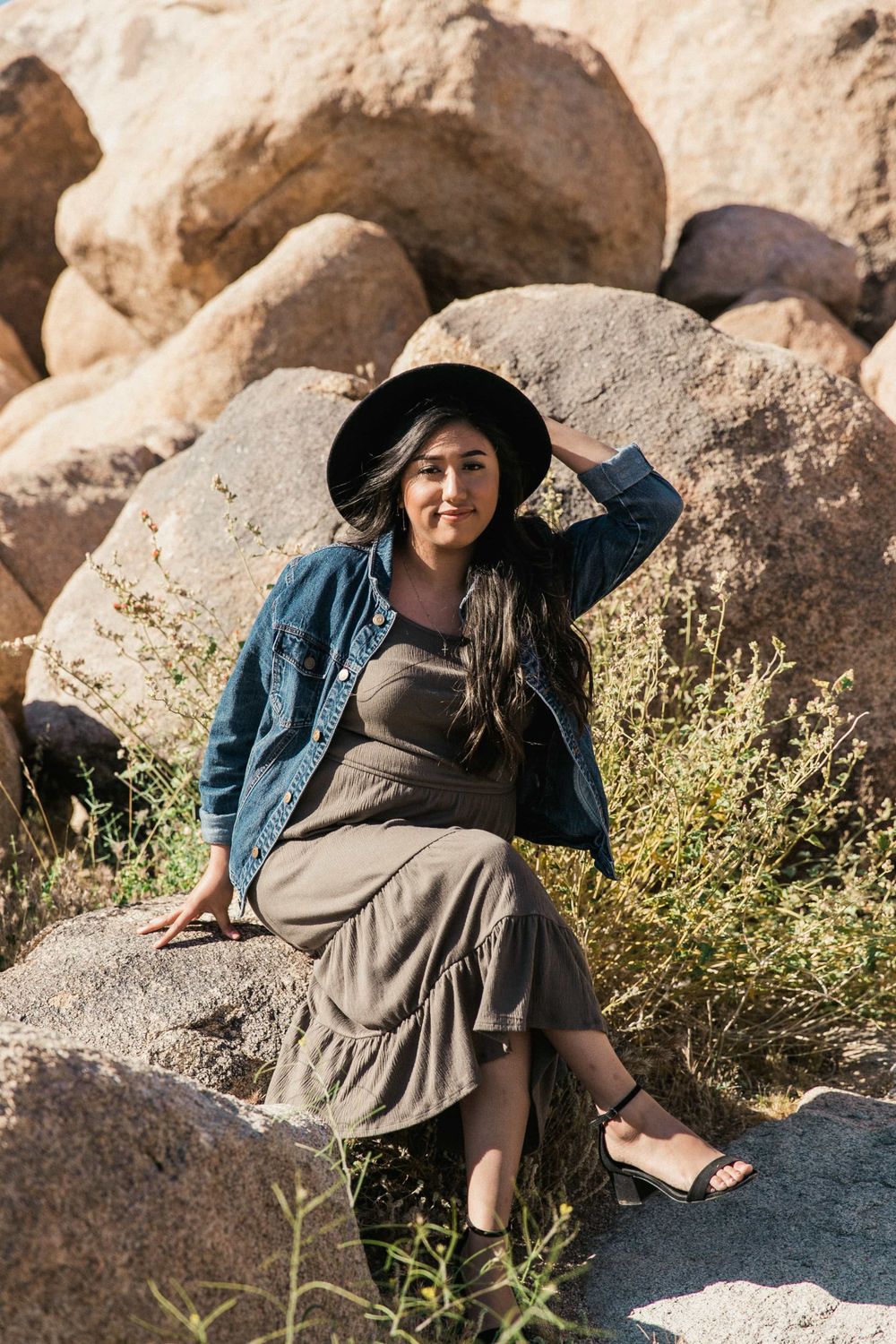 Smiling woman in gray dress, denim jacket, and black hat sits relaxed on rocks in sunny desert setting.