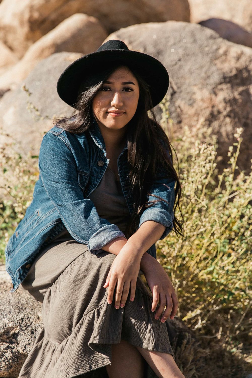 Young woman in black hat and denim jacket sits thoughtfully on a rock amid desert boulders and wildflowers.