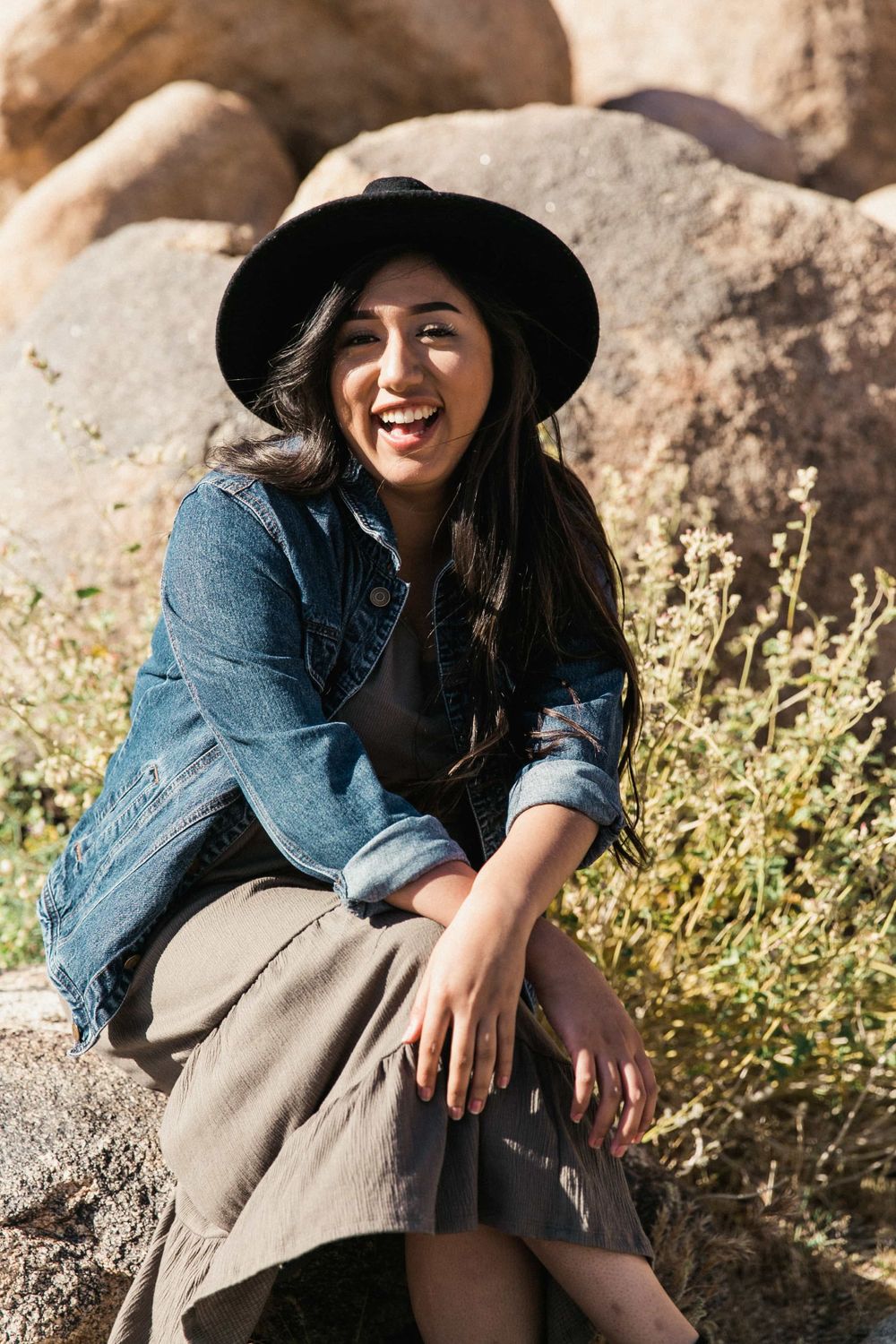 Laughing woman in black hat and denim jacket sits joyfully on a rock in a sunny desert landscape.