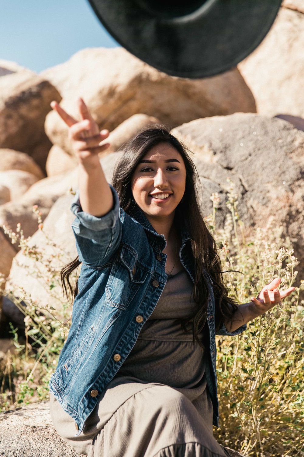Cheerful young woman in denim jacket flashes a peace sign while sitting among desert boulders outdoors.