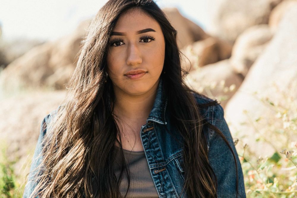 Close-up portrait of a young woman with long dark hair in a denim jacket among desert boulders.