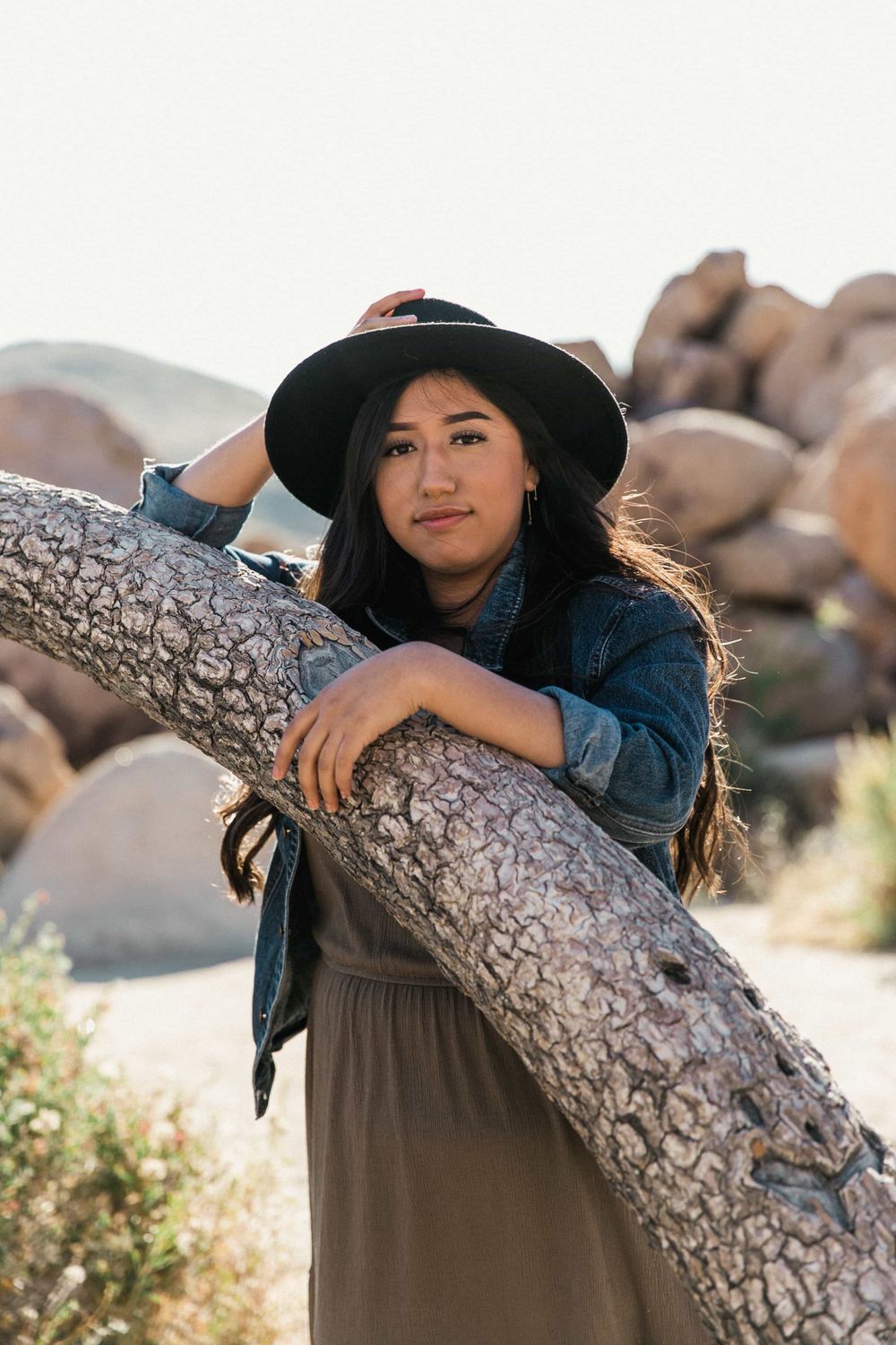 Young woman in black hat and denim jacket leaning on a tree trunk in a rocky desert landscape.