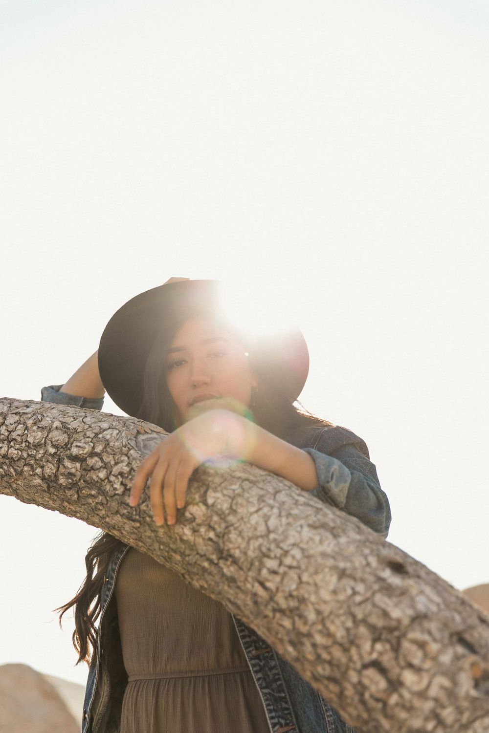 Woman in black hat leaning on a tree trunk with sun flare in a bright desert setting.
