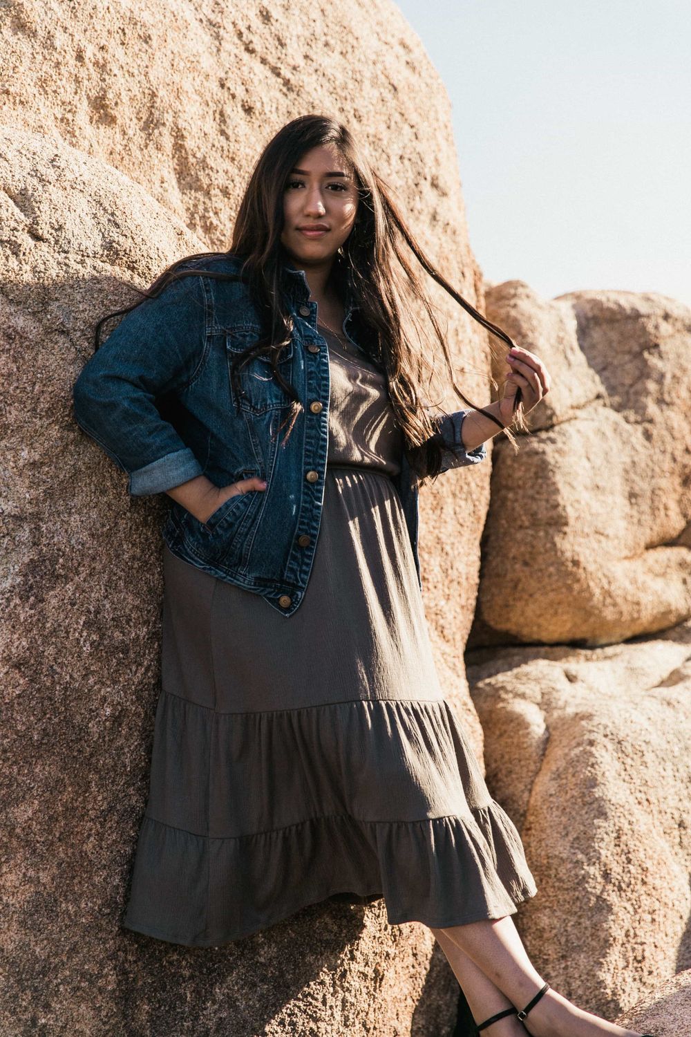 Young woman in denim jacket and olive maxi dress posing against large boulders outdoors.