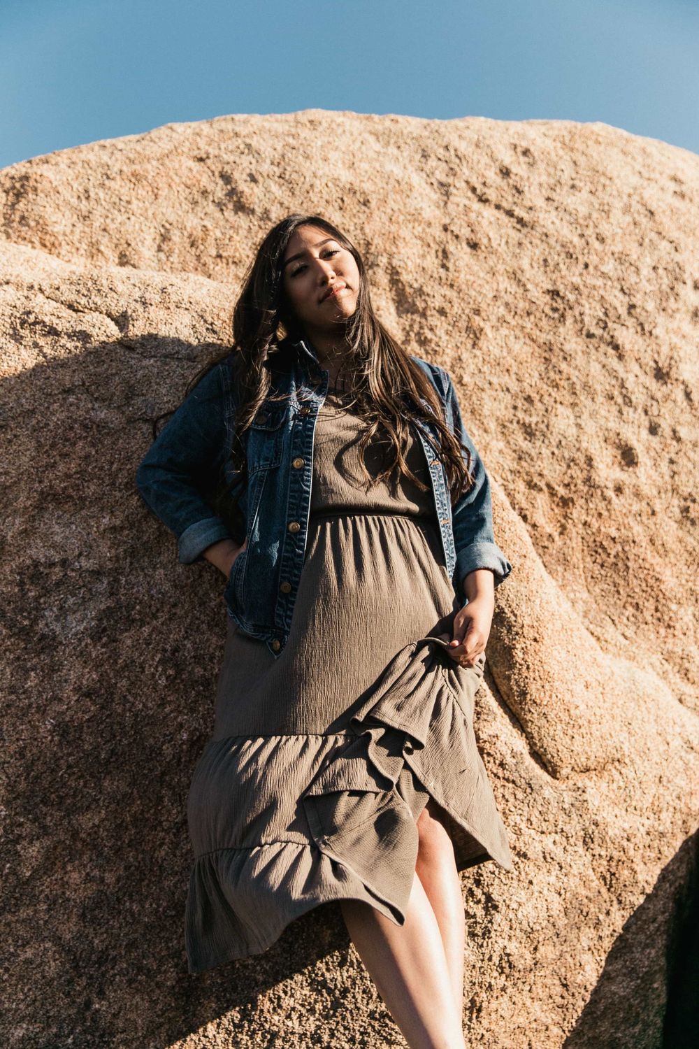 Woman in denim jacket and olive dress leaning against a large boulder under a blue sky.