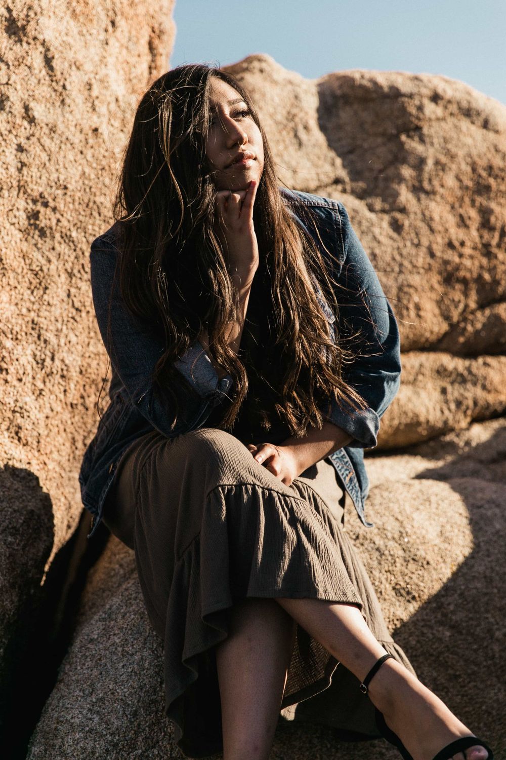 Young woman in denim jacket and olive skirt sitting on rocks in a sunny desert setting.