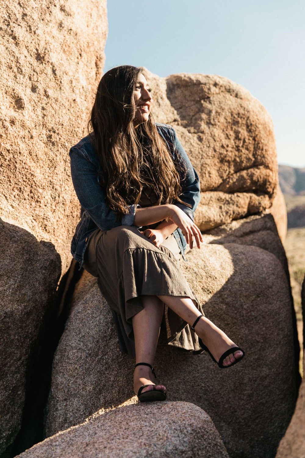 Young woman in denim jacket and olive skirt sitting on rocks smiling in a sunny desert.