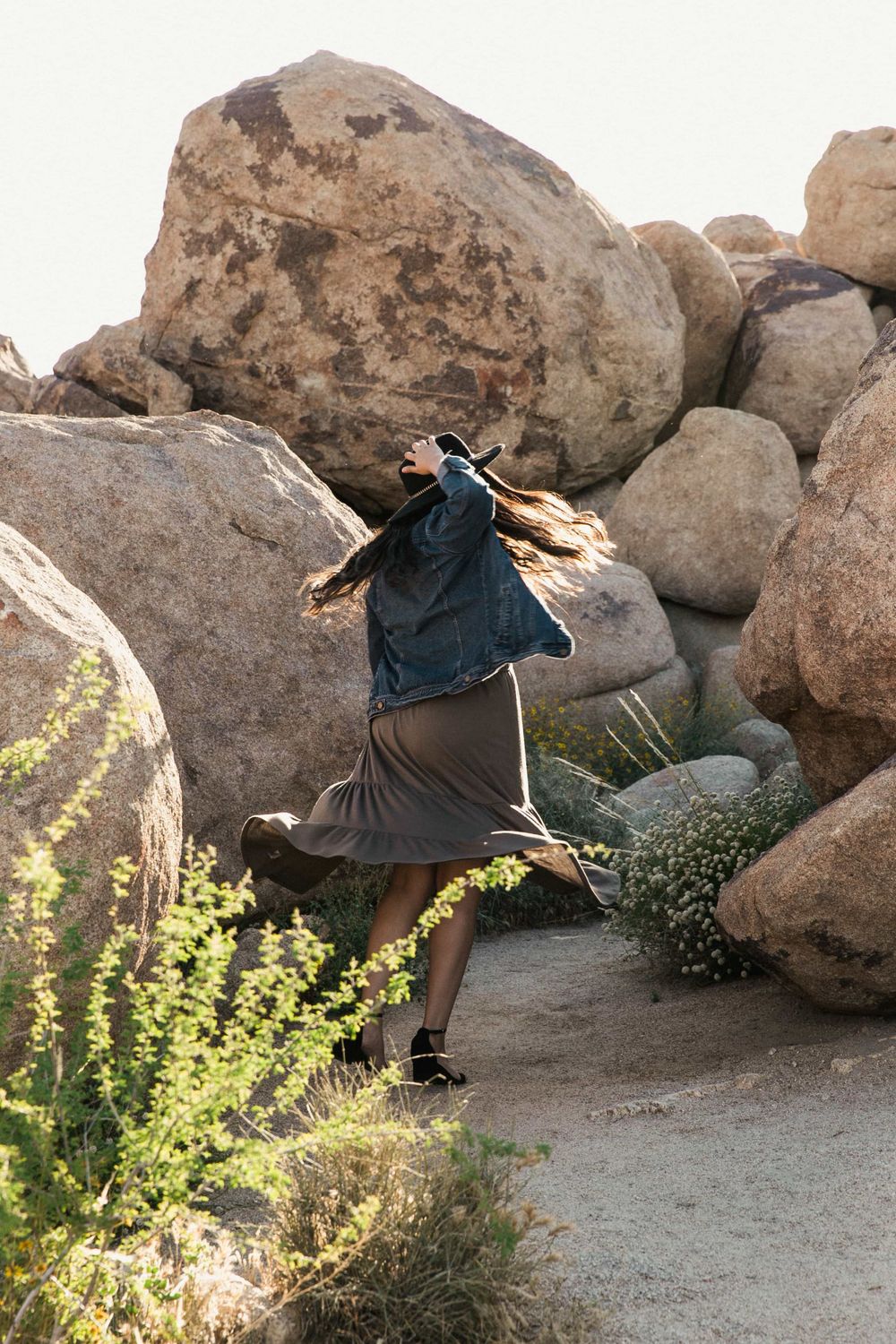 Woman in denim jacket and olive skirt walking among large boulders in a desert landscape.