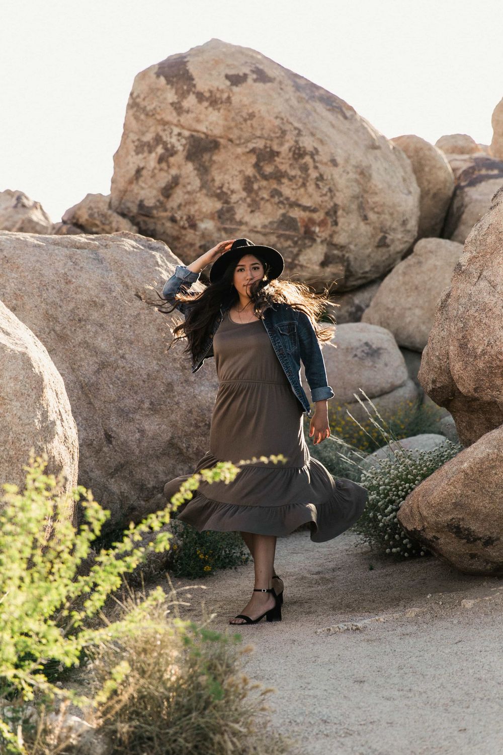 Woman in black hat and denim jacket twirling in an olive dress among desert boulders.