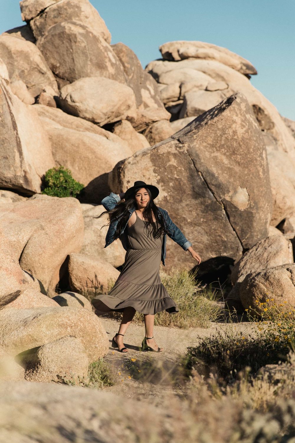 Woman in black hat and denim jacket walking in an olive dress among large desert boulders.