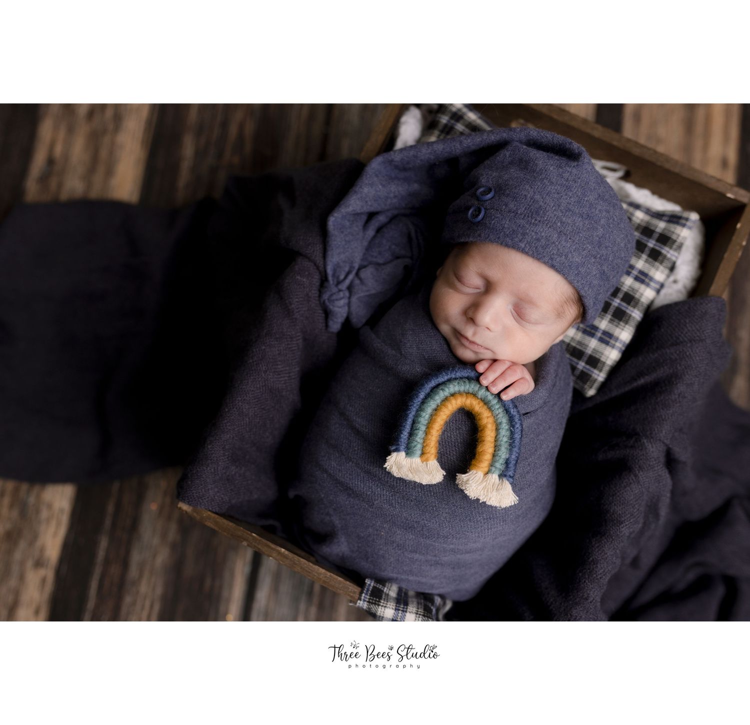 Newborn in navy outfit with rainbow design sleeping on plaid pillow.