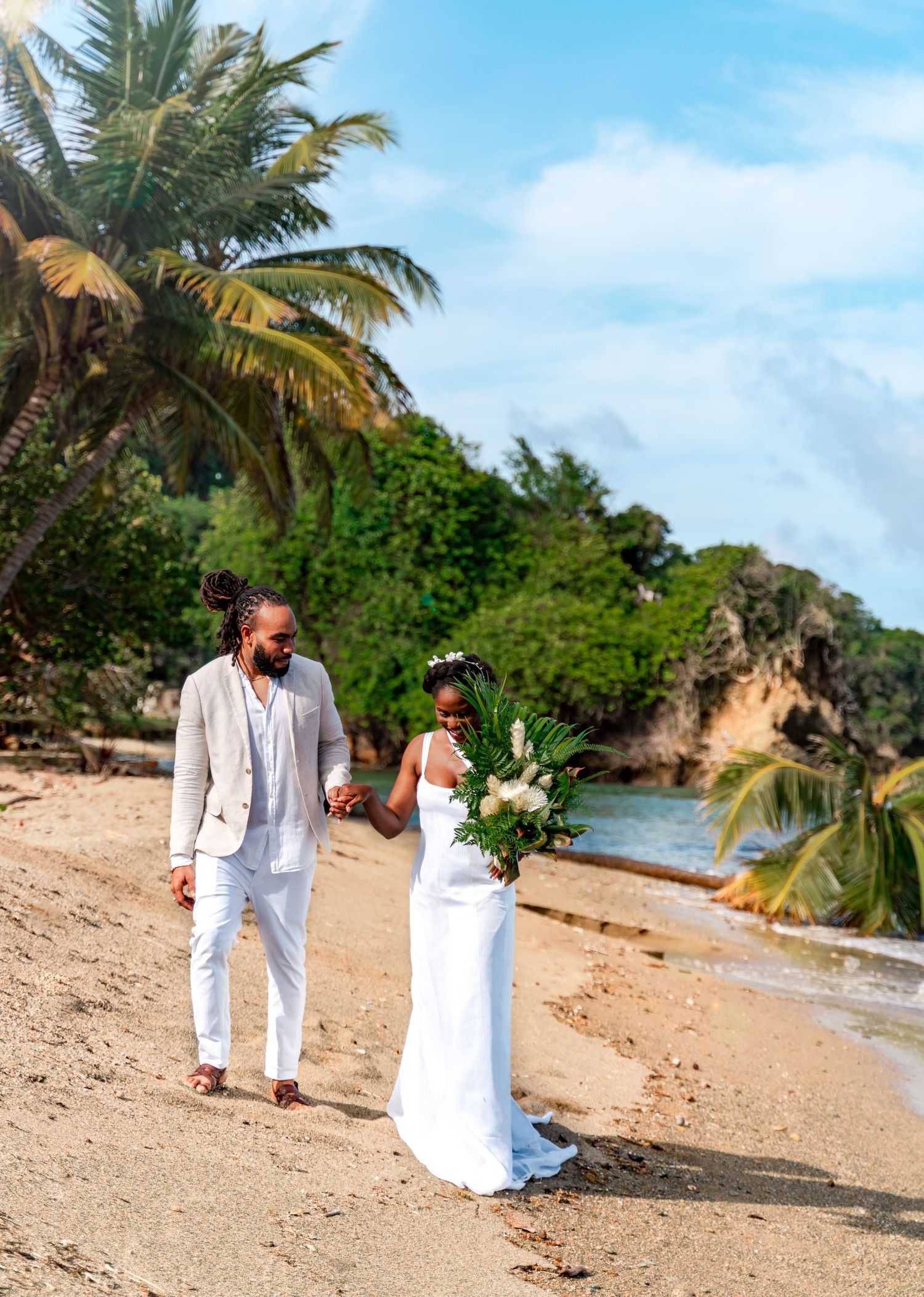 A beach wedding couple walking along a palm-lined tropical shoreline in white attire.