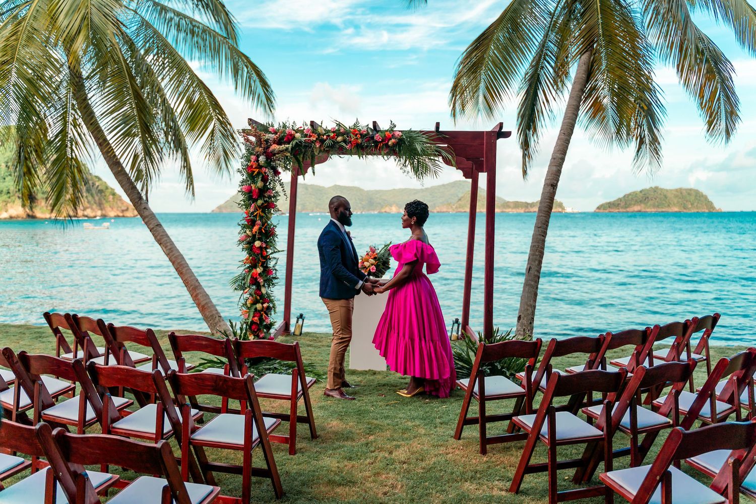 A tropical beach wedding ceremony setup with pink flowers and wooden chairs under palm trees.