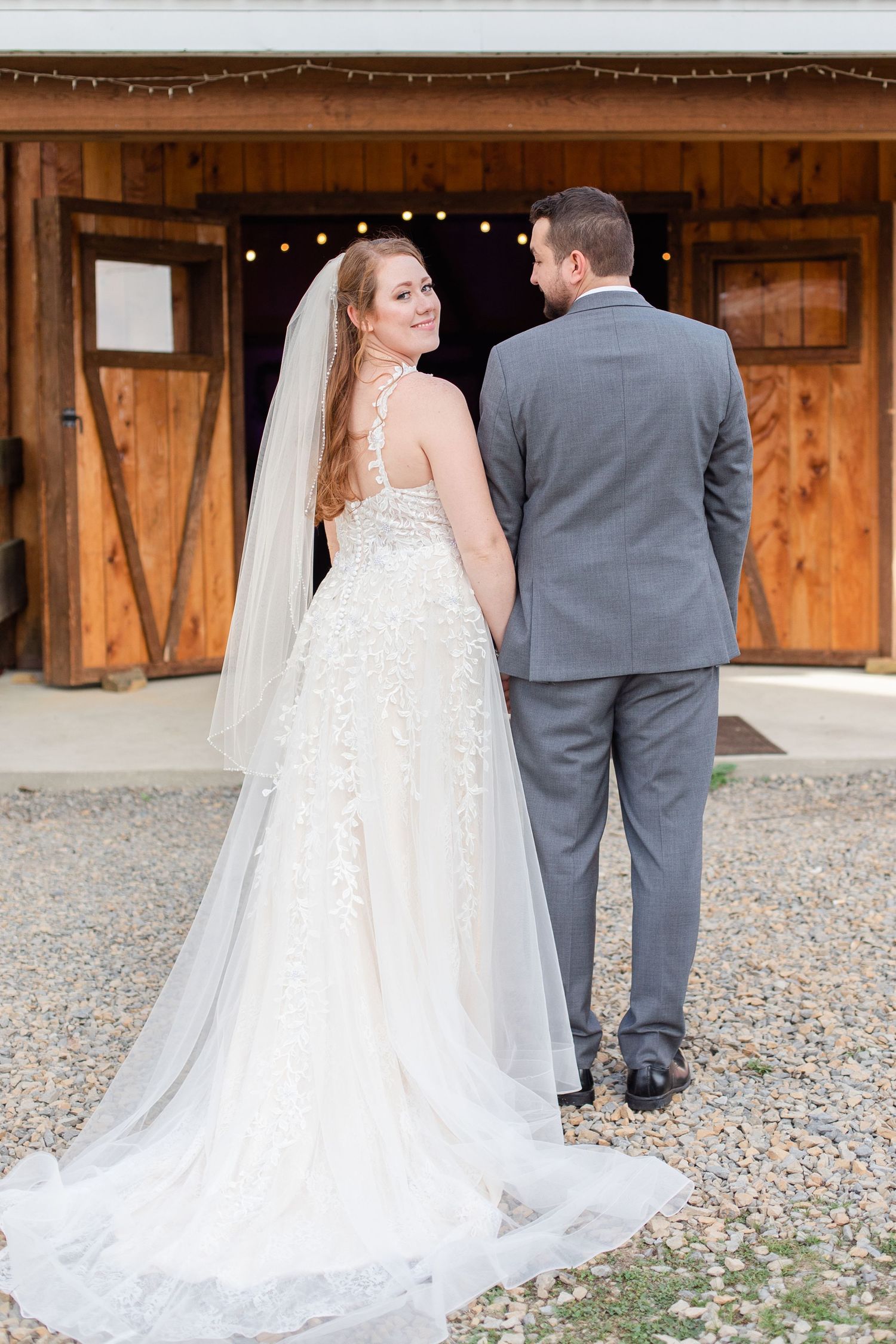Wedding couple poses near rustic barn showing details of bridal gown train.