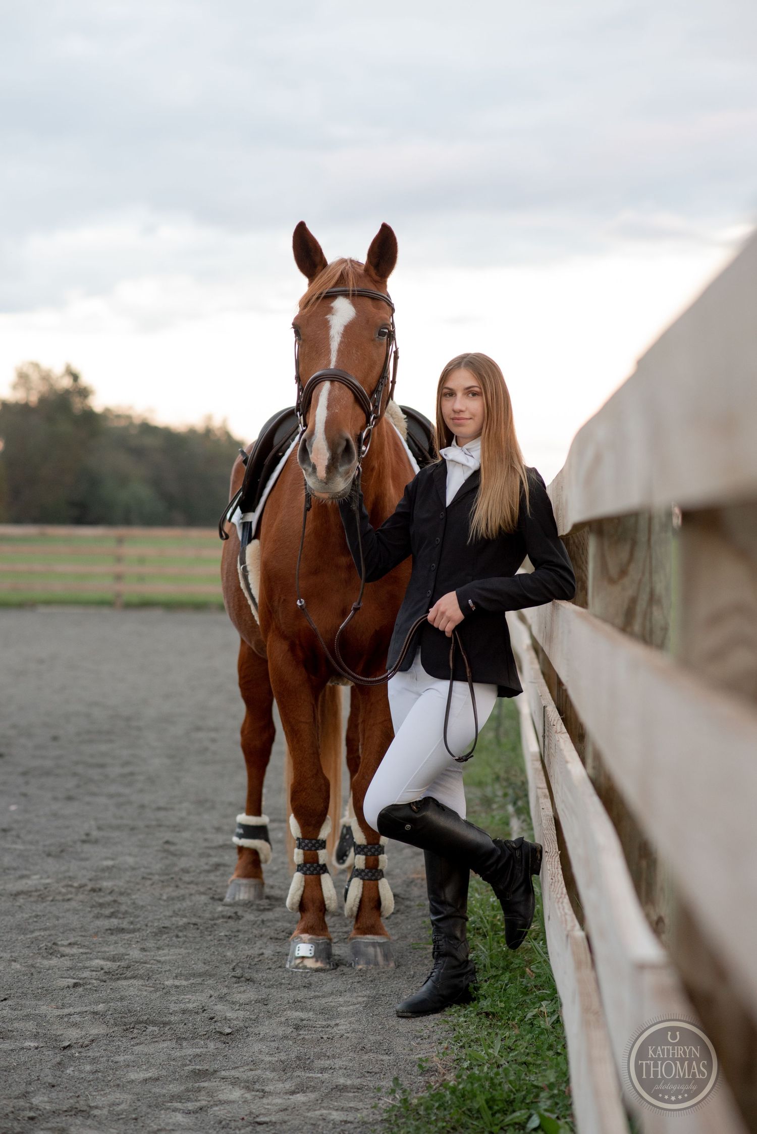 Equestrian in riding attire poses with saddled horse along arena fence.