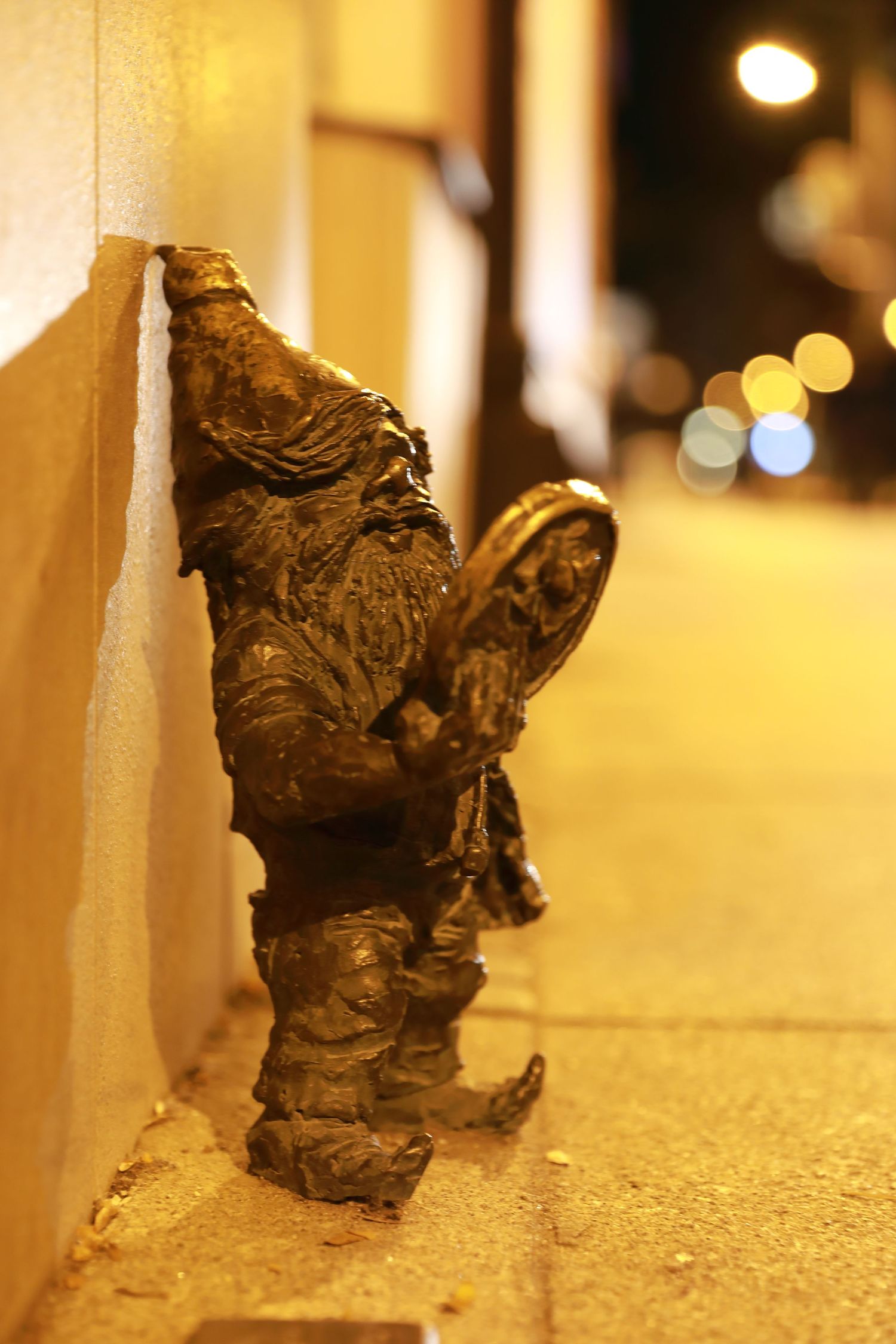 Bronze gnome statue against wall with bokeh lights in background.