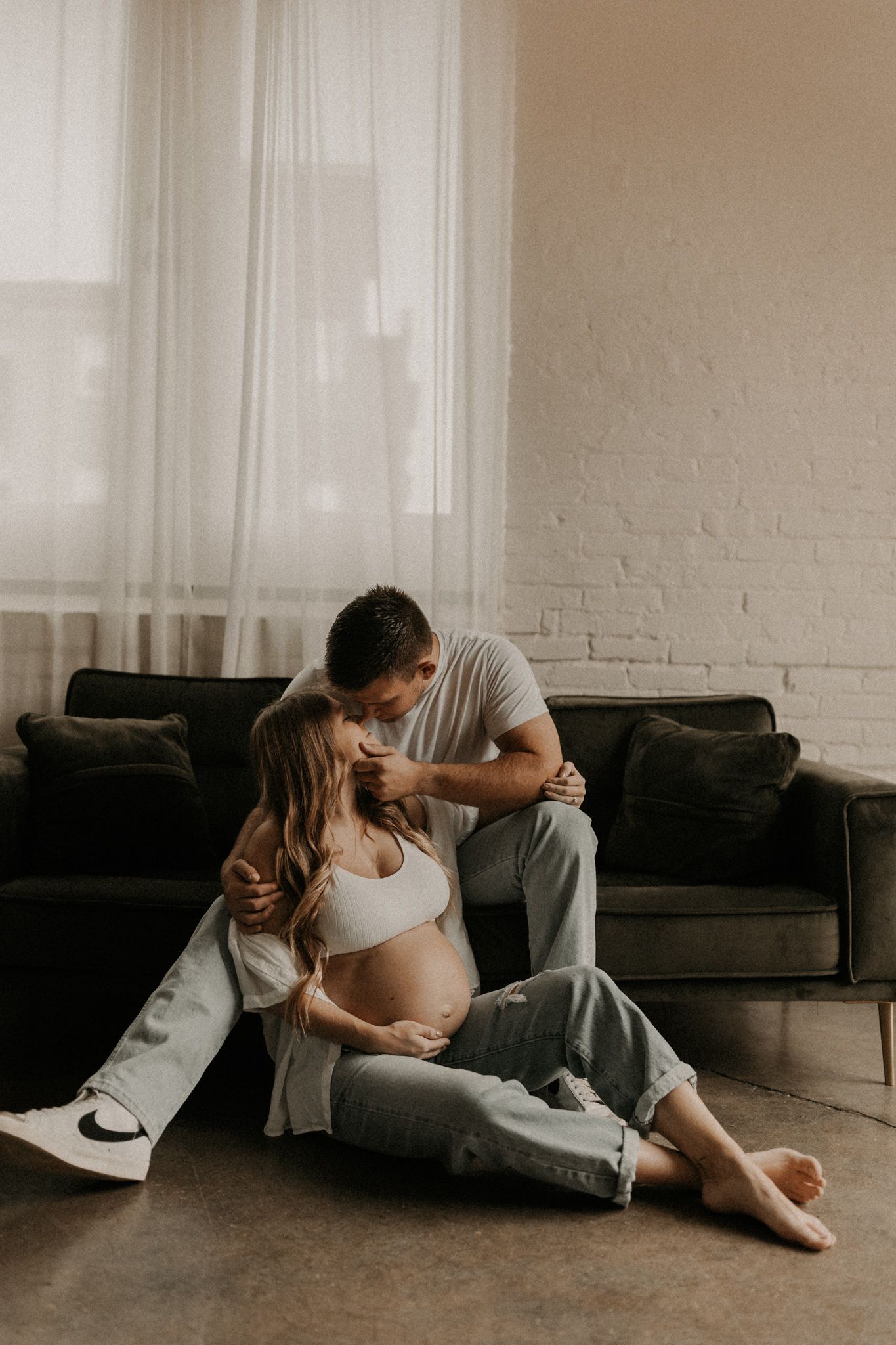 An intimate maternity photo session with a couple on a dark velvet couch in a minimalist setting.