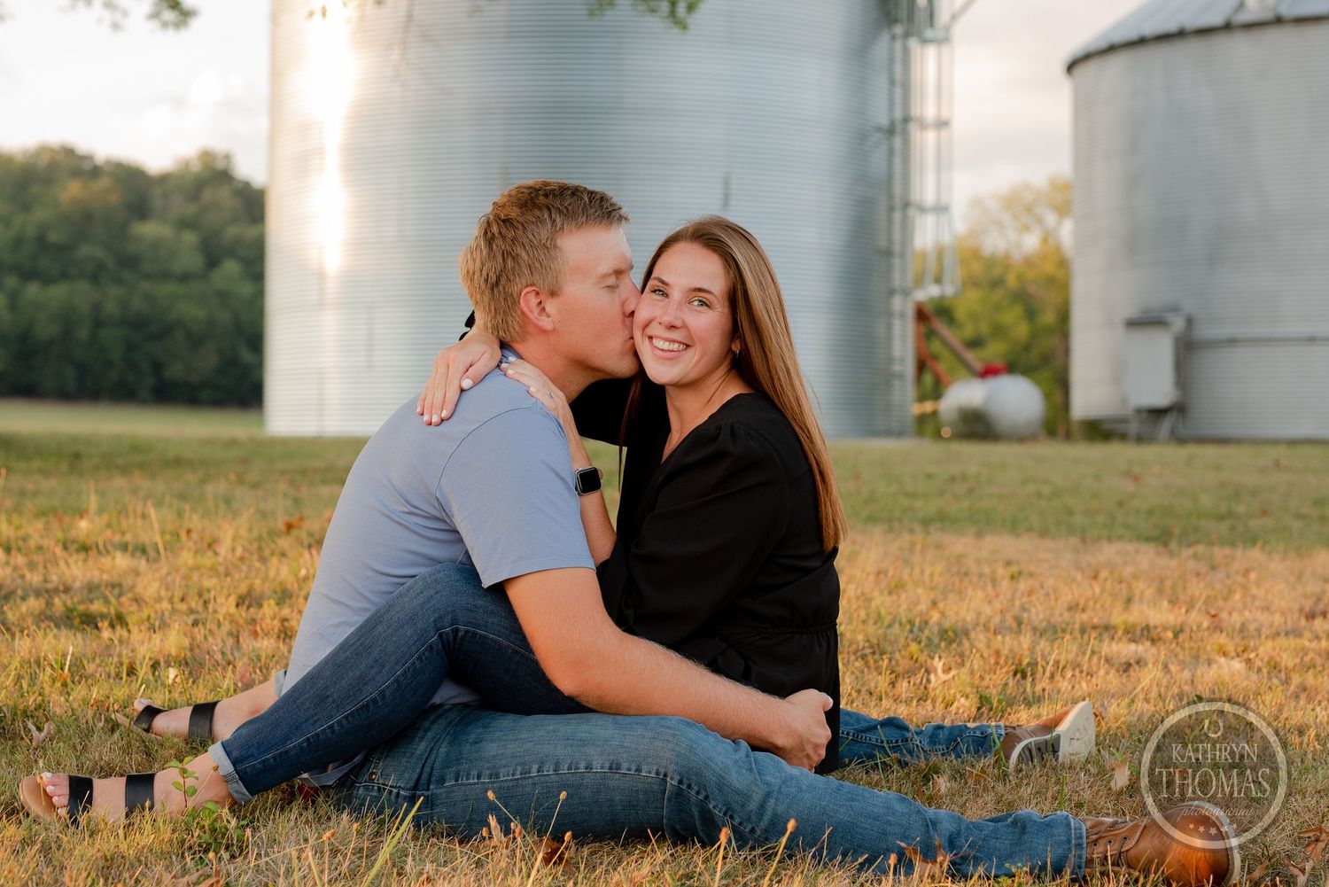 Couple sitting together on grass with industrial silos in background at sunset.