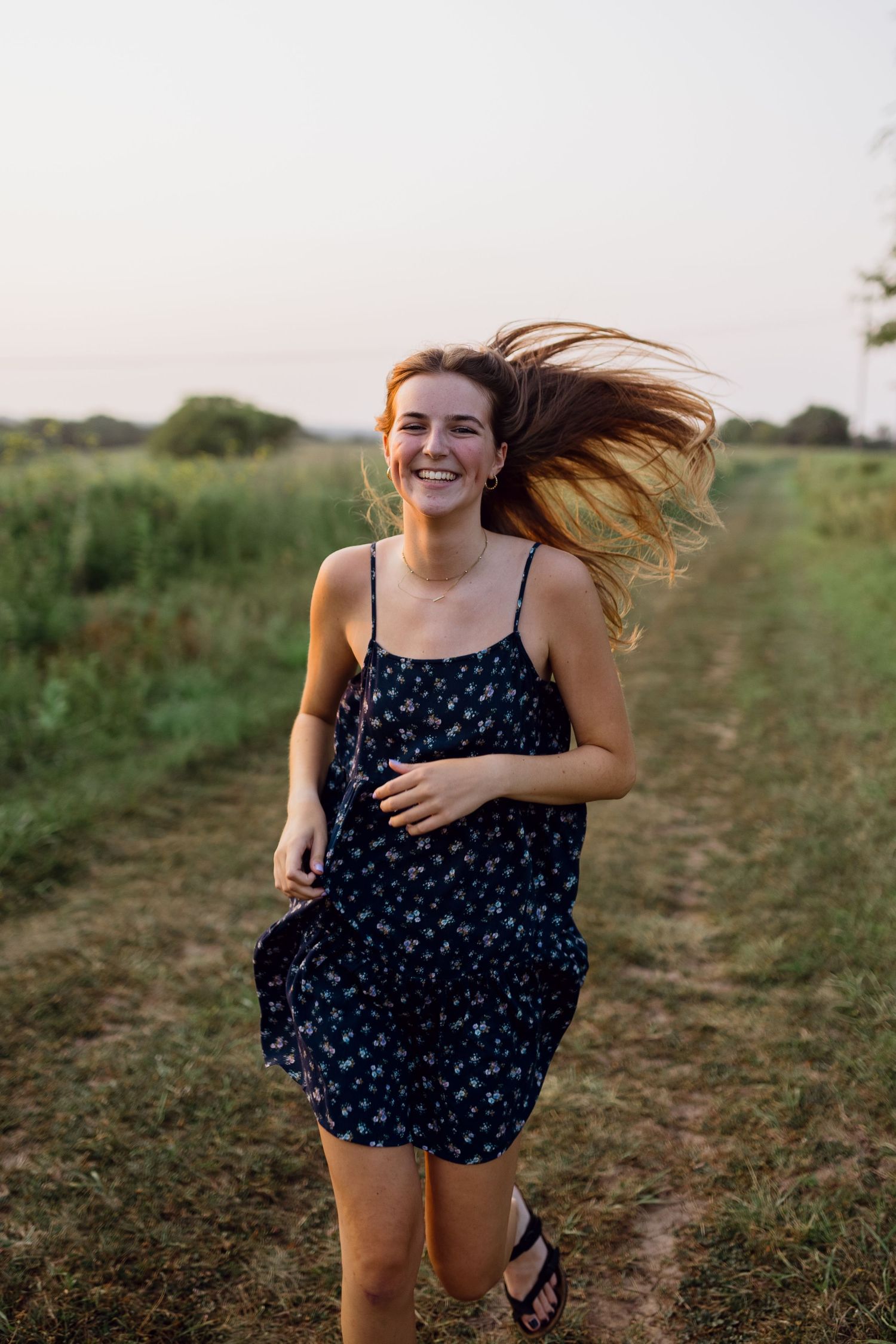 A high school senior in a floral dress runs with wind in her hair through an open field during a senior portrait session