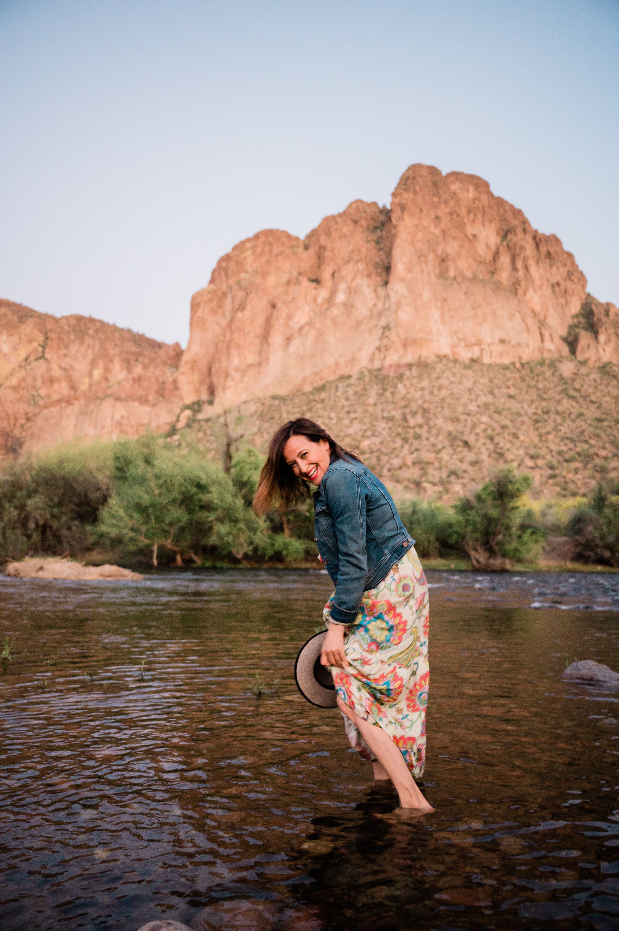 Someone in a denim jacket and floral dress plays in the water beneath towering desert cliffs.