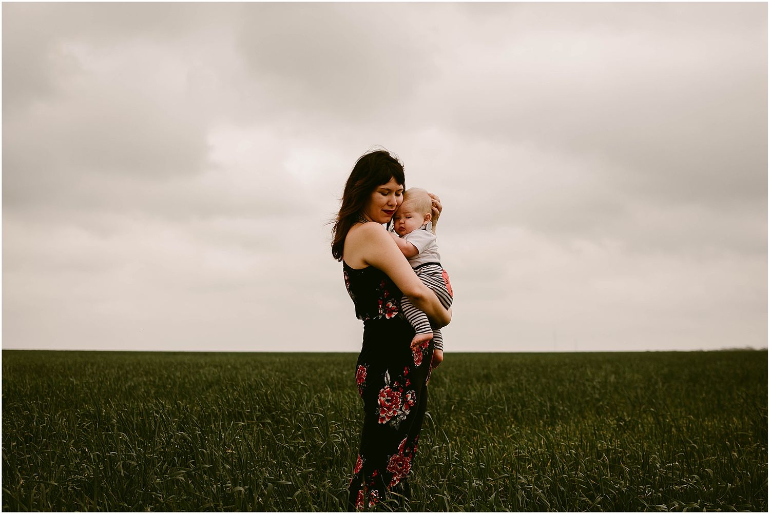 Silhouette of adult holding baby against cloudy sky in open meadow landscape.