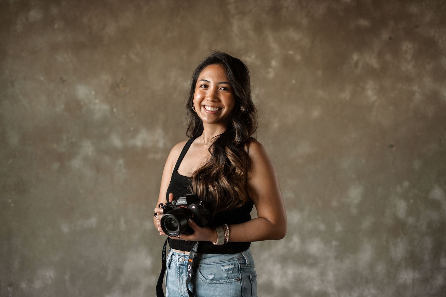 Woman in casual denim and black top poses with a professional camera against a neutral wall.