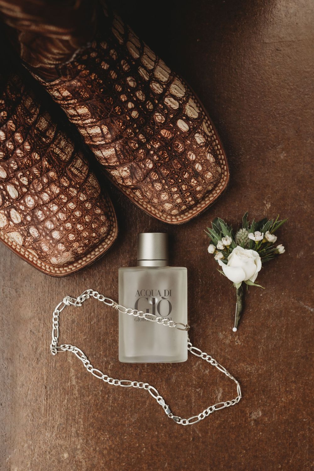 Wedding accessories including leather boots, perfume bottle, chain necklace and boutonniere on brown surface.