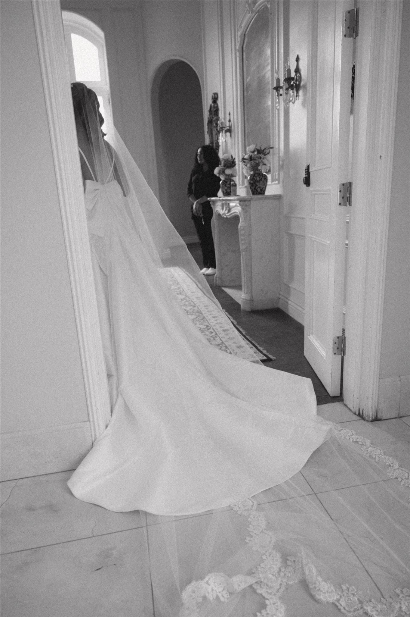 A dramatic black and white photograph shows a flowing wedding gown with a long lace train in an elegant white hallway.