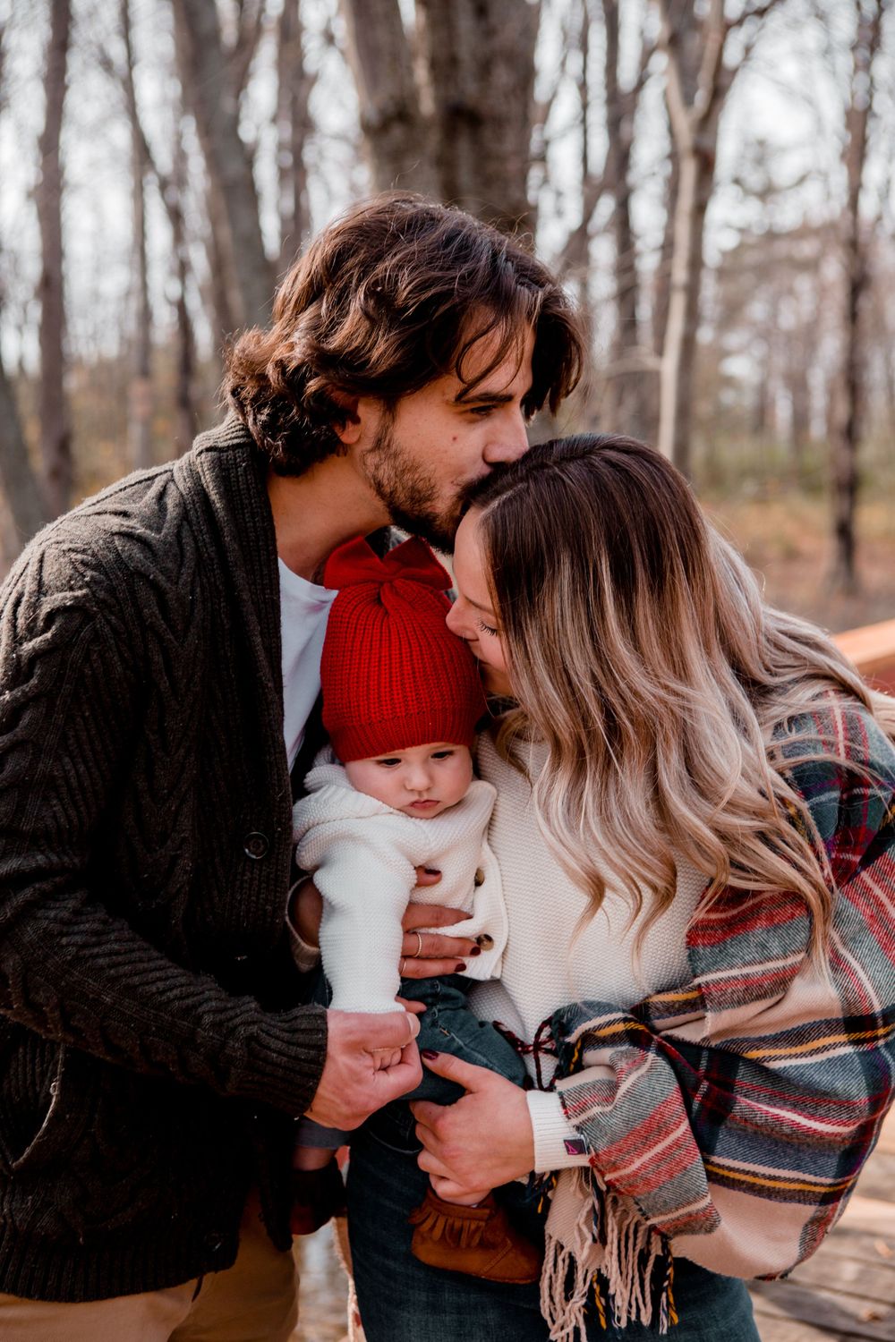 A cozy autumn family moment in the woods with a baby wearing a red hat.