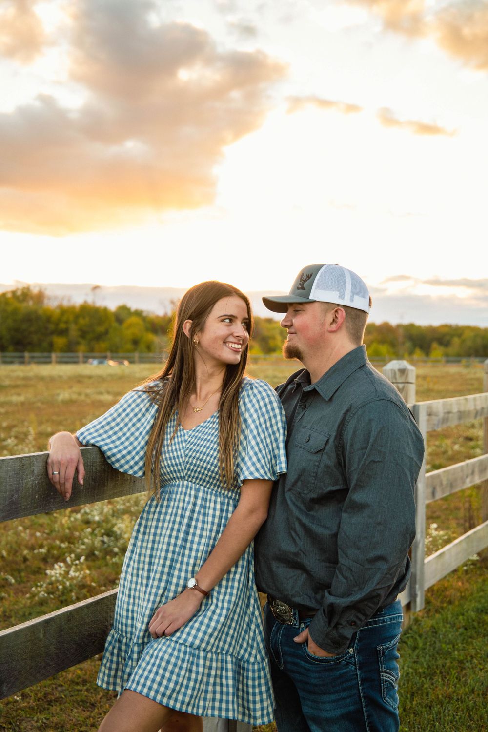 Couple sharing intimate moment in field during sunset with rustic fence backdrop.
