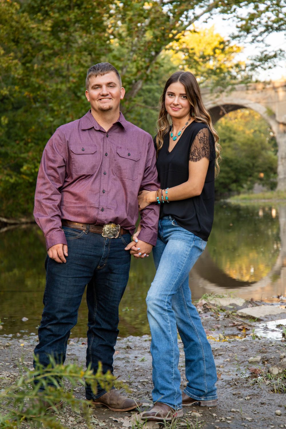 Couple poses by a scenic pond with fall foliage and stone bridge in background.