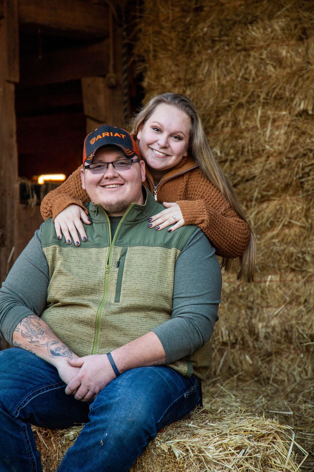Couple cuddling in a rustic barn setting with hay bales in the background.