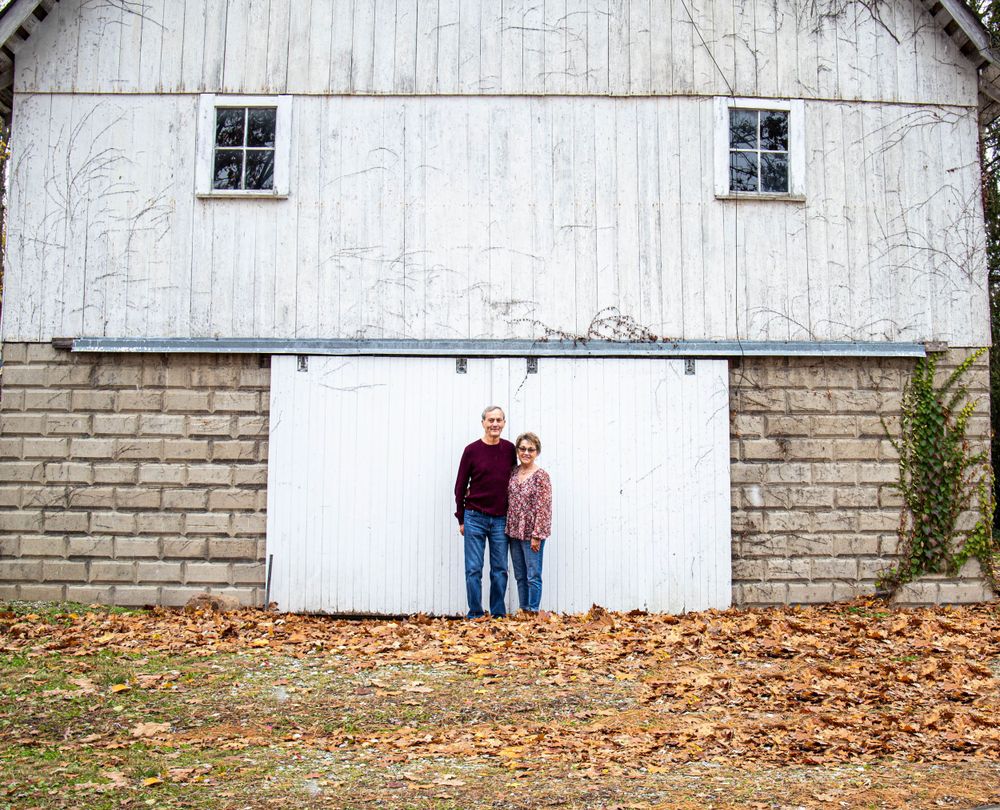 Couple poses in front of rustic white barn with blue door during fall season.