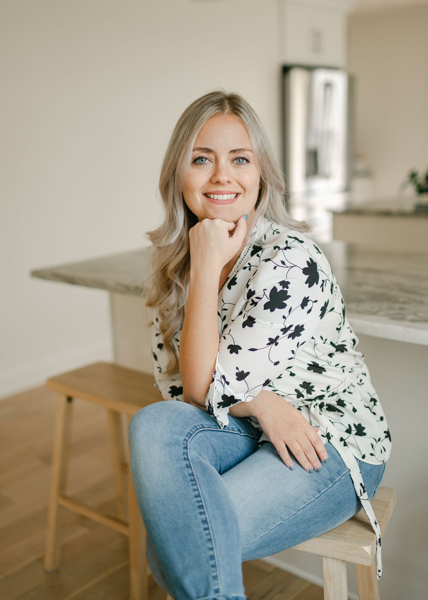 Person in floral print top seated on wooden stool in bright modern kitchen interior.
