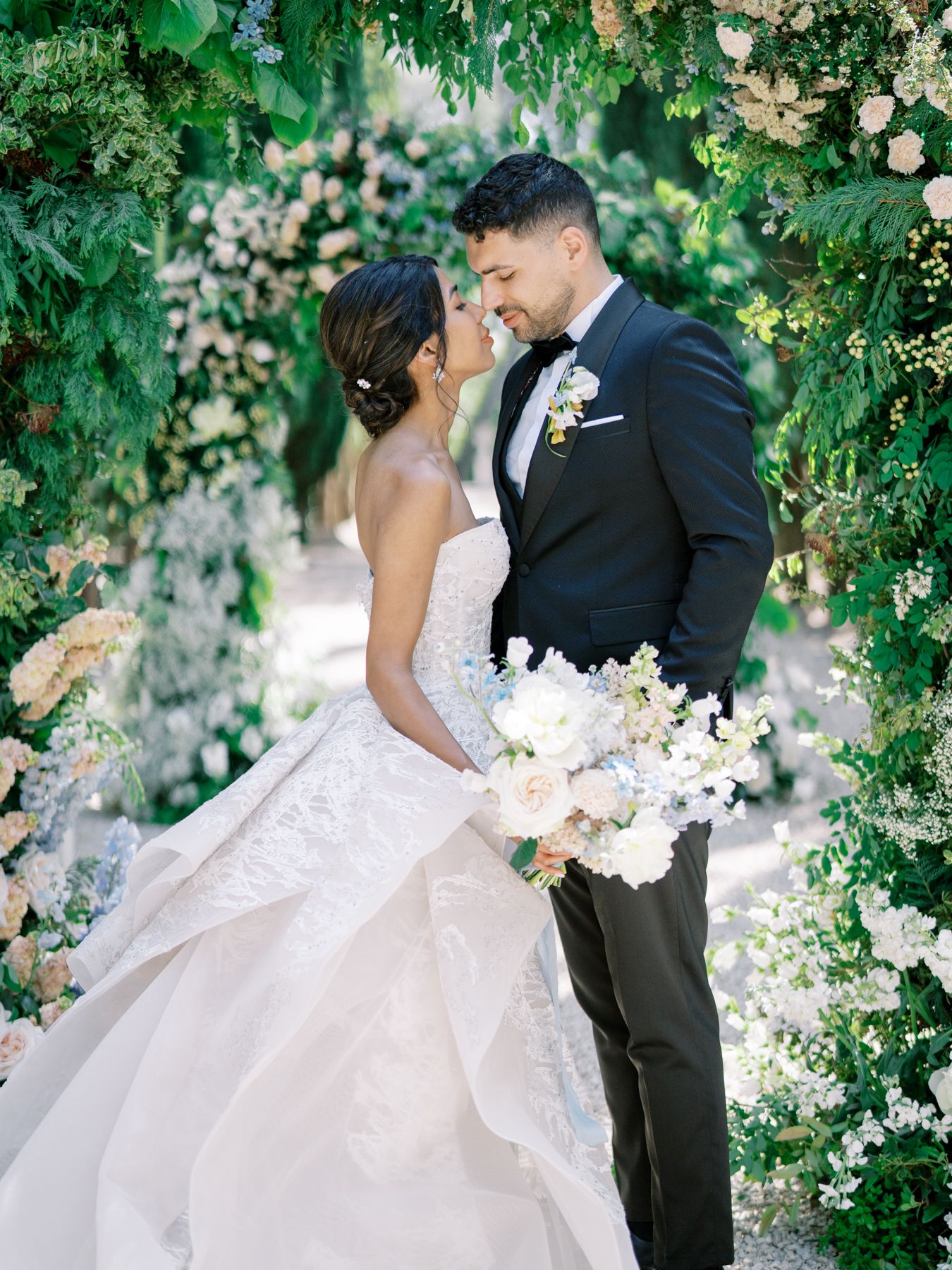 Elegant wedding couple embracing in a garden archway surrounded by white flowers.