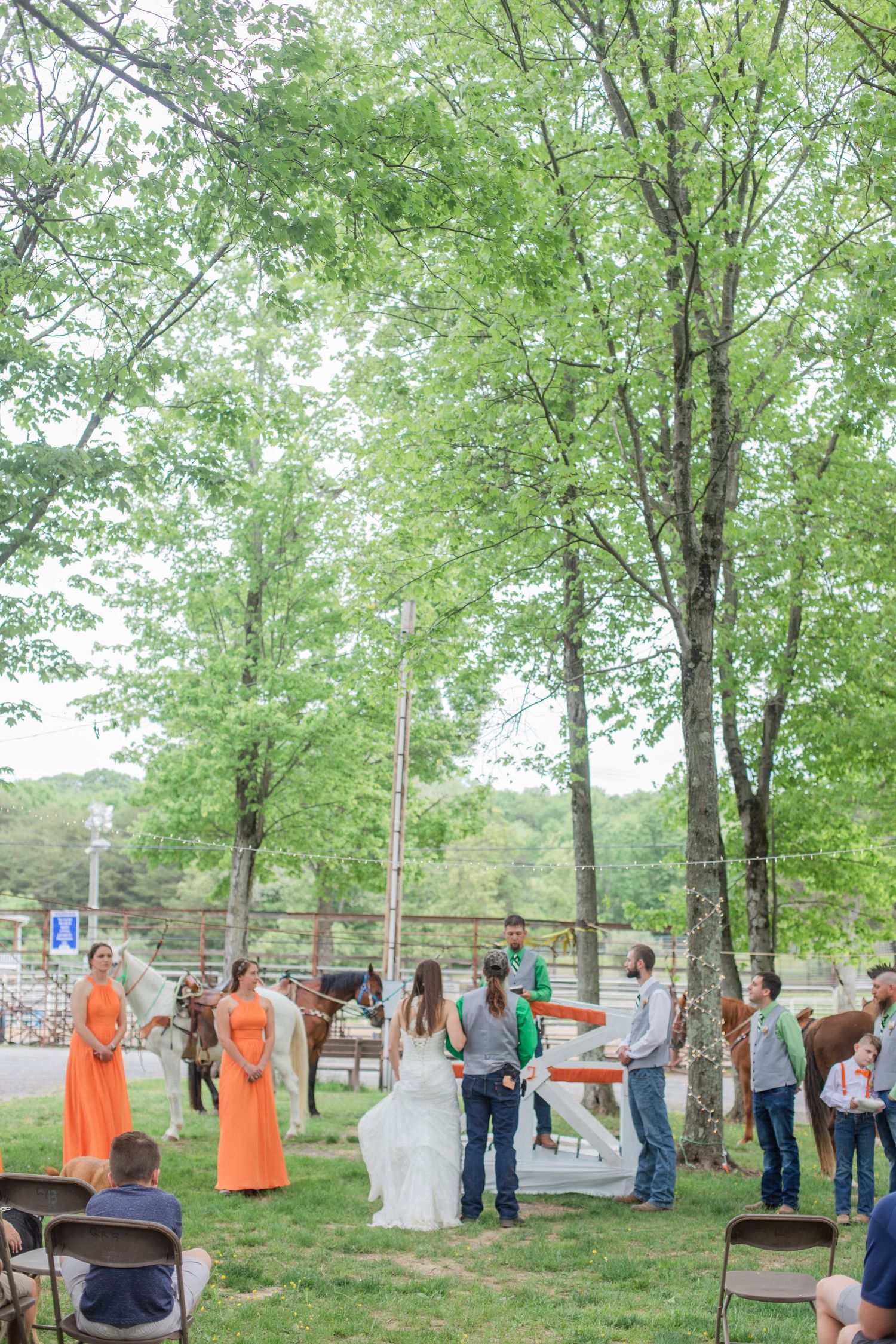An outdoor wedding gathering under tall green trees with orange-clad bridesmaids and casual guests.
