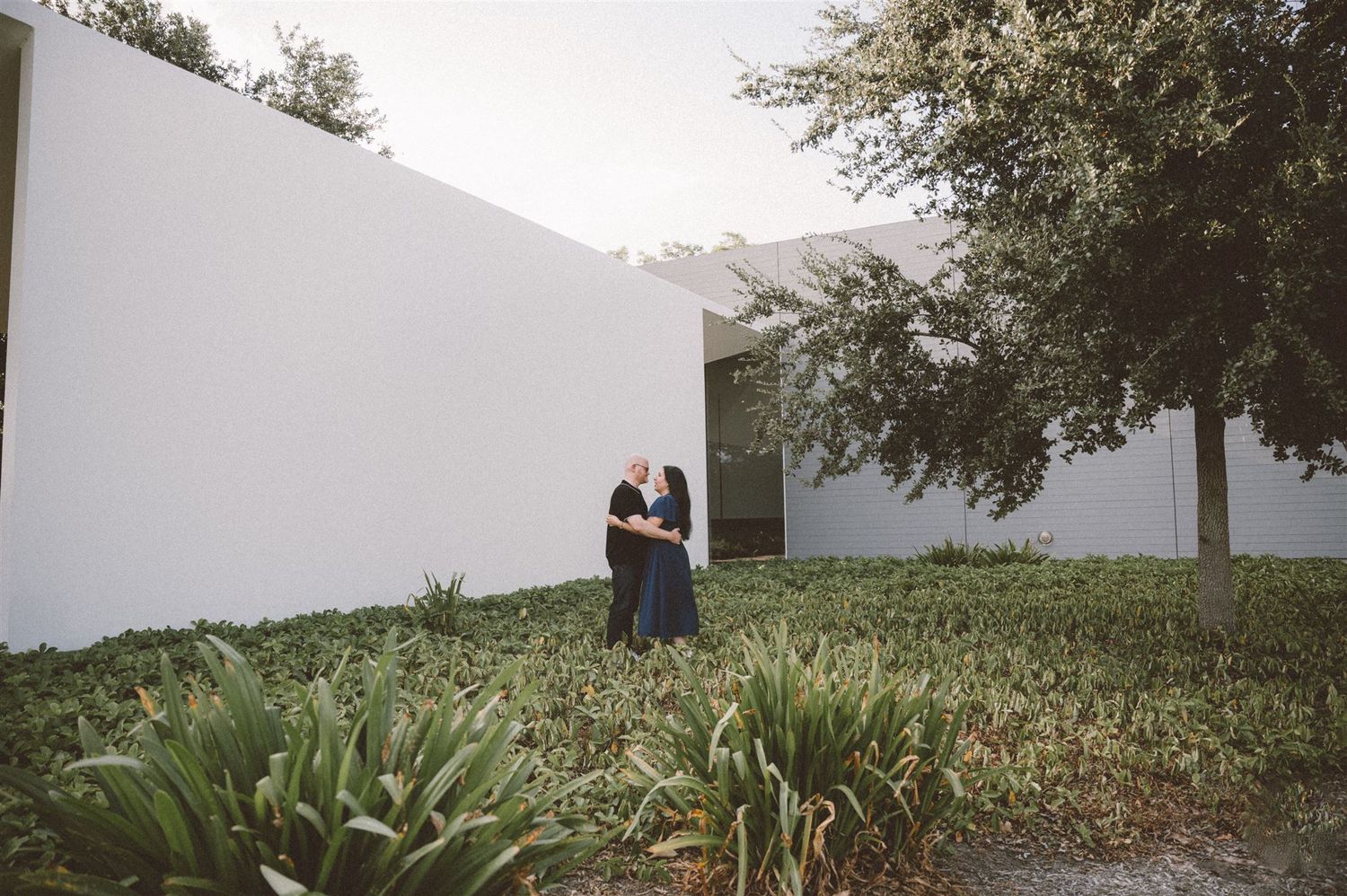 A couple embraces on a grassy hill against white architectural walls.