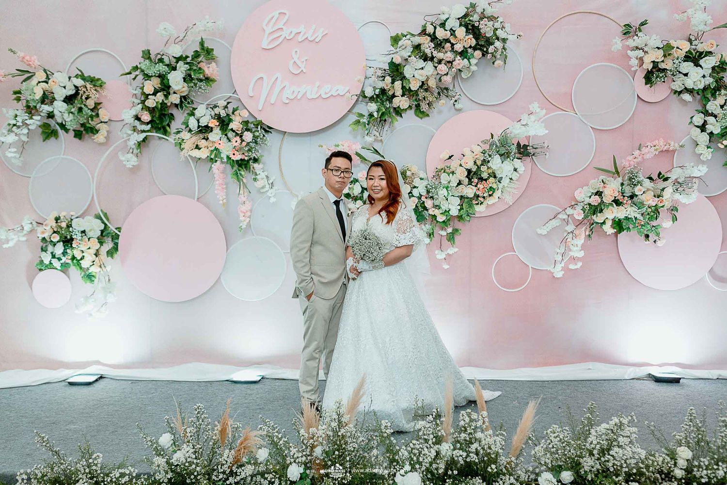 Wedding couple posing in front of elegant pink and white circular backdrop with floral arrangements.