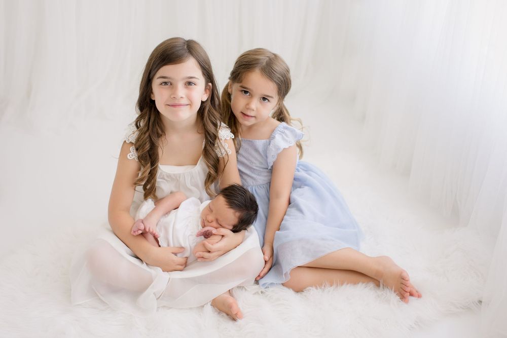 Three siblings in soft blue dresses posed together with newborn on white background.