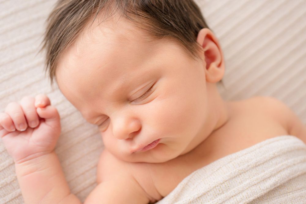 Peaceful sleeping newborn wrapped in soft white fabric against a neutral background.