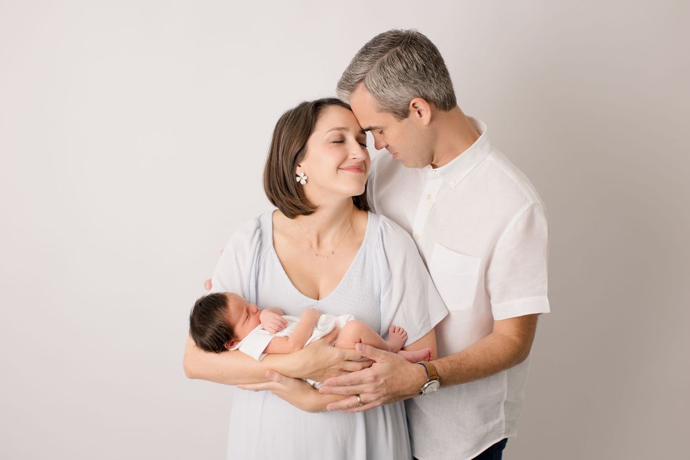 Intimate family portrait with parents holding newborn baby against neutral background.